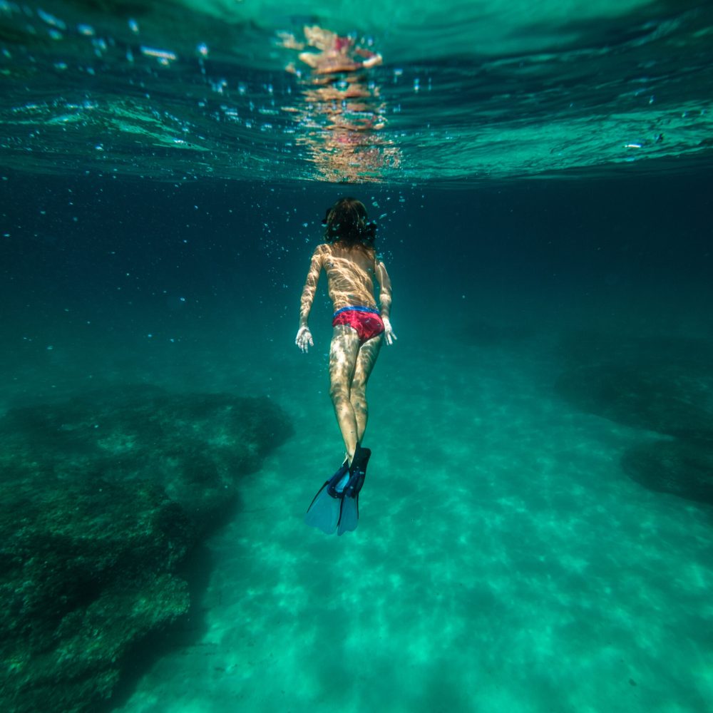 Back view of full body anonymous kid traveler in diving mask and flippers swimming underwater and enjoying beautiful coral reef and undersea world