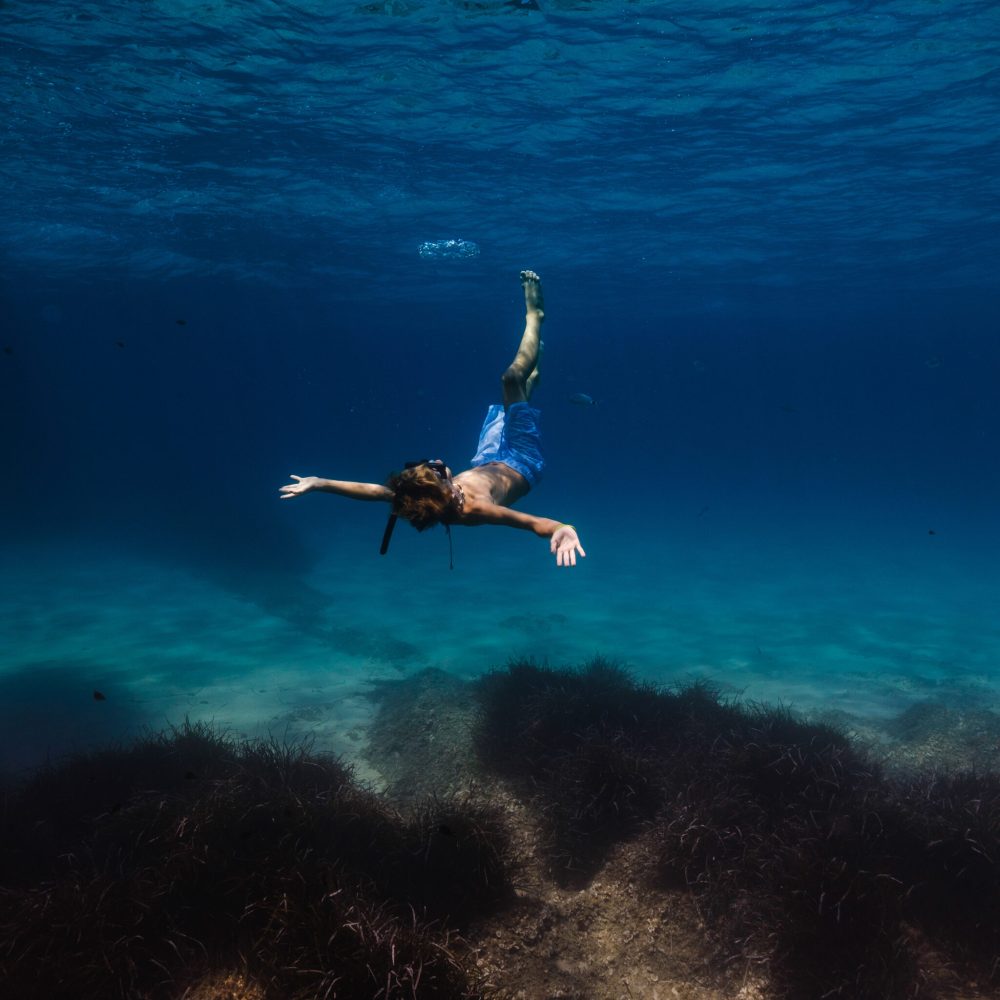 Full body of unrecognizable kid in snorkeling mask and tube floating under blue seawater with corals on sandy bottom and fish