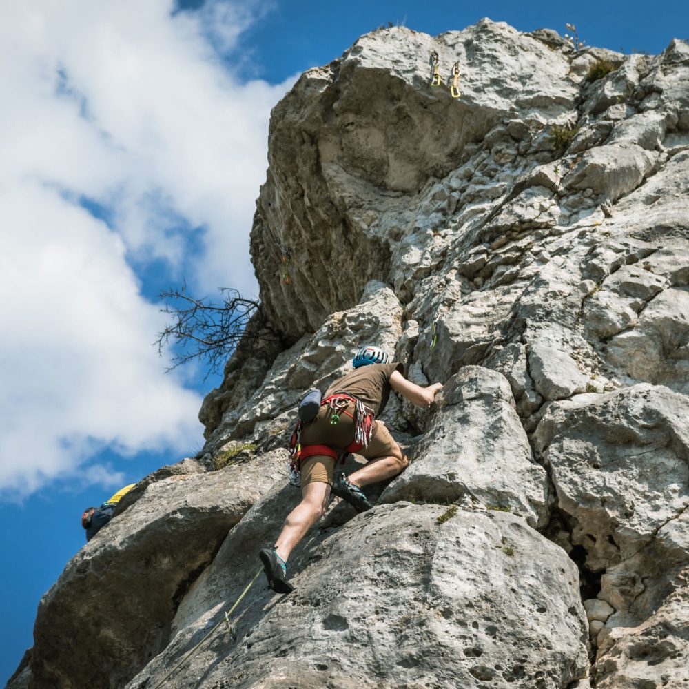 A low-angle rear of a man climbing a high rock, cloudy nd sunlit sky background