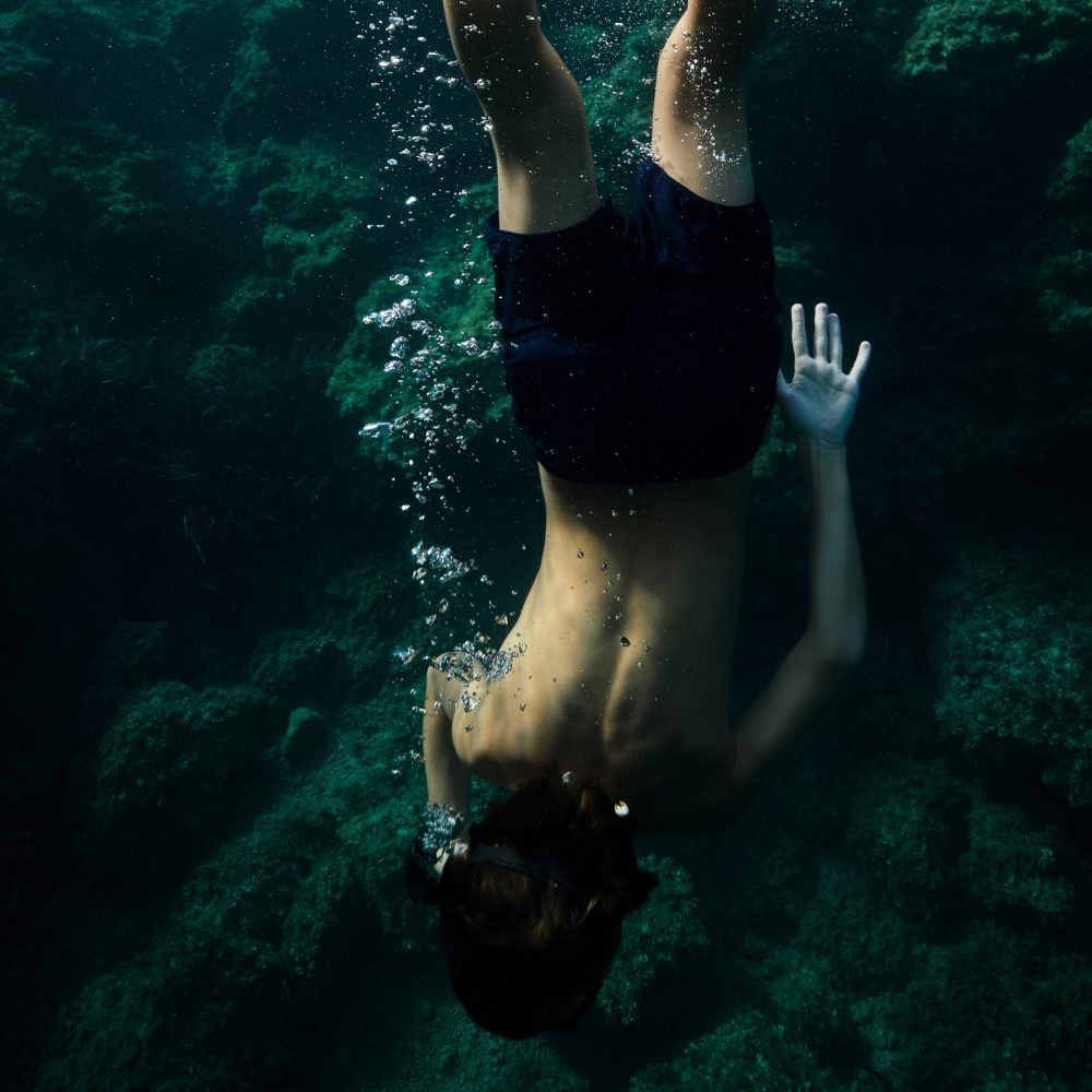 Back view full body of anonymous male diver in wetsuit and flippers diving under transparent seawater with coral reefs on bottom